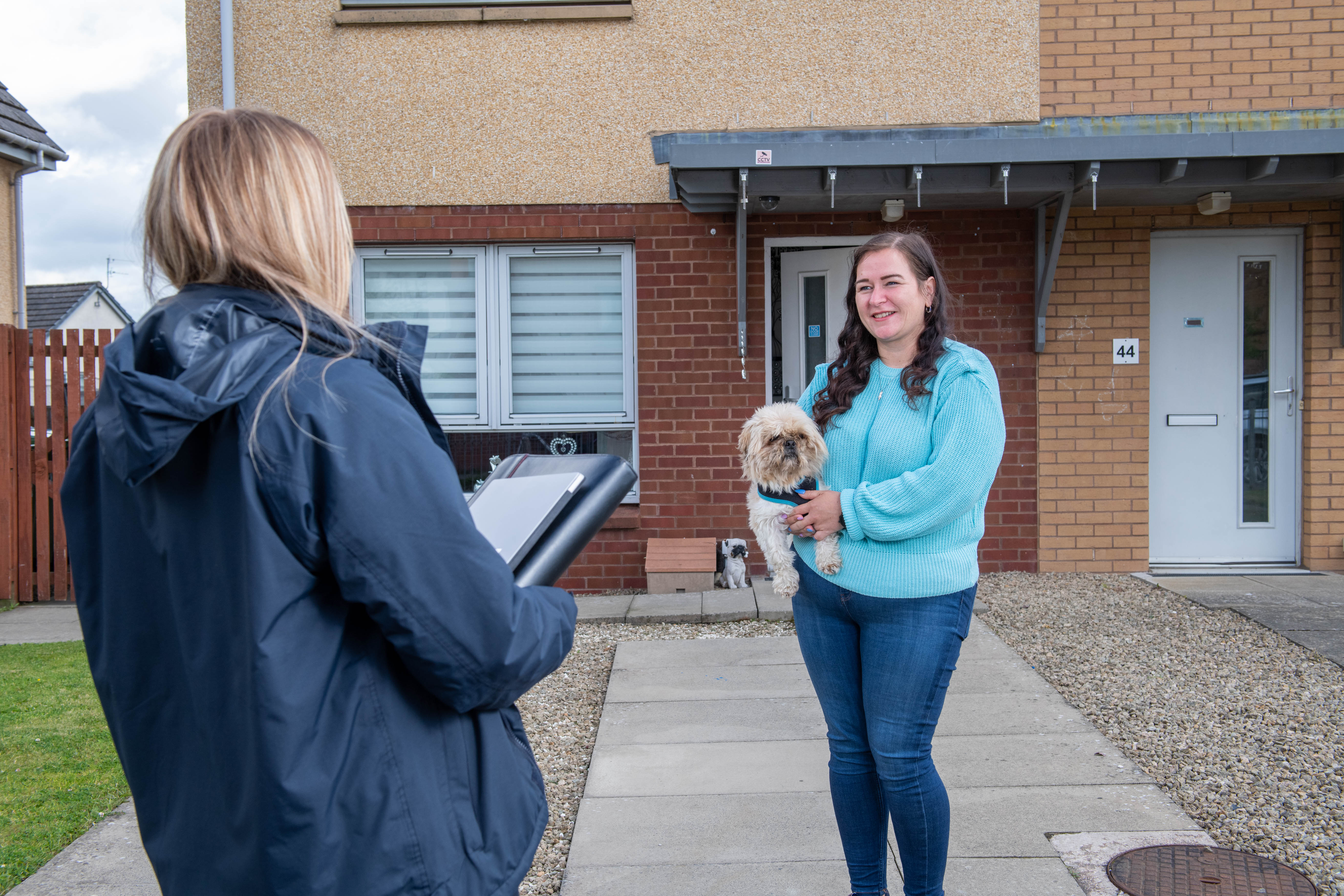 Who we say we are news image A person outside the front of their home holding a small dog facing another person who looks as though they are working for an organisation with a folder in their hand