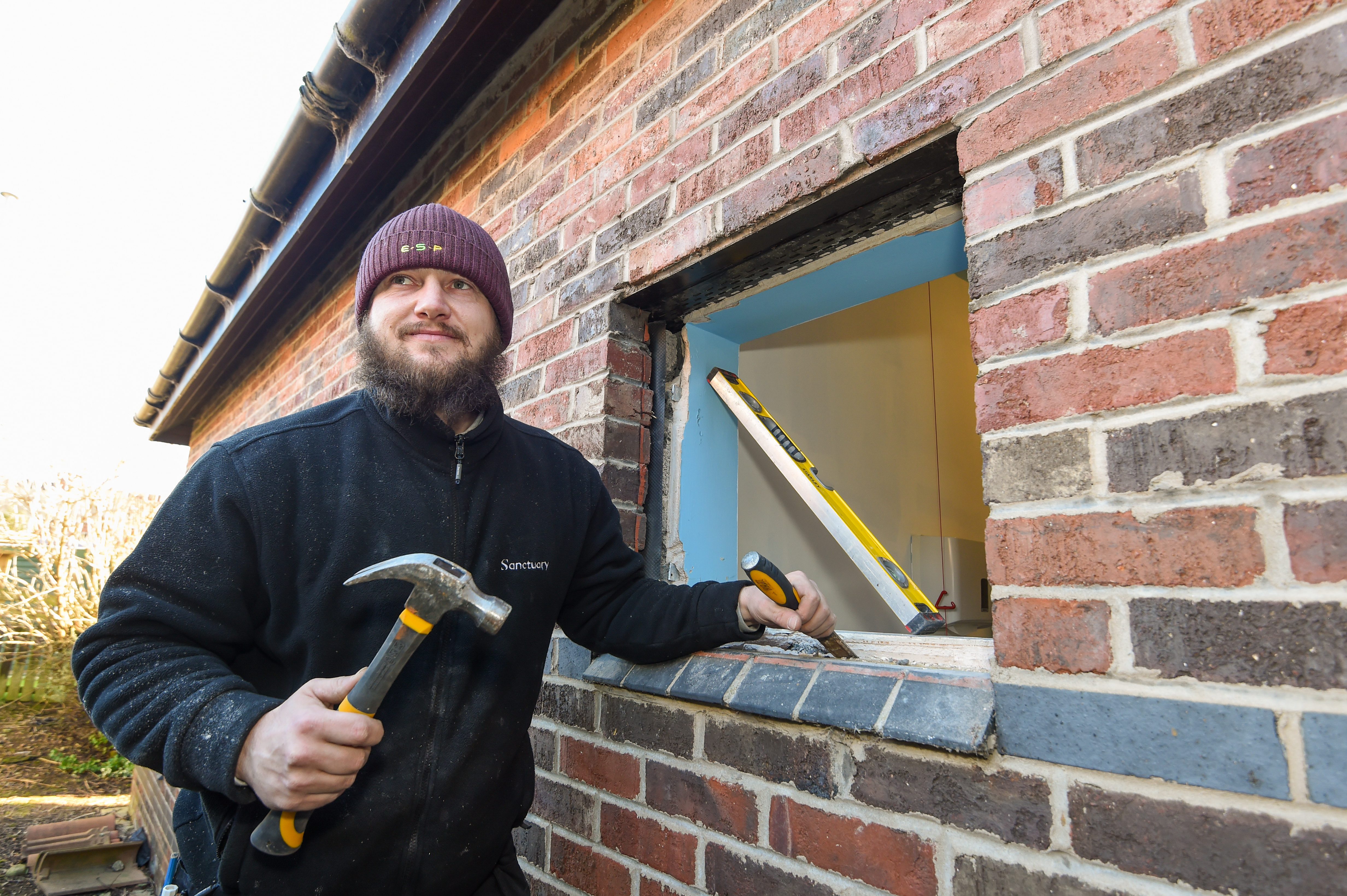 Replacing window A man in a navy Sanctuary-branded fleece holds a chisel and hammer in front of a window where the double-glazing has been removed