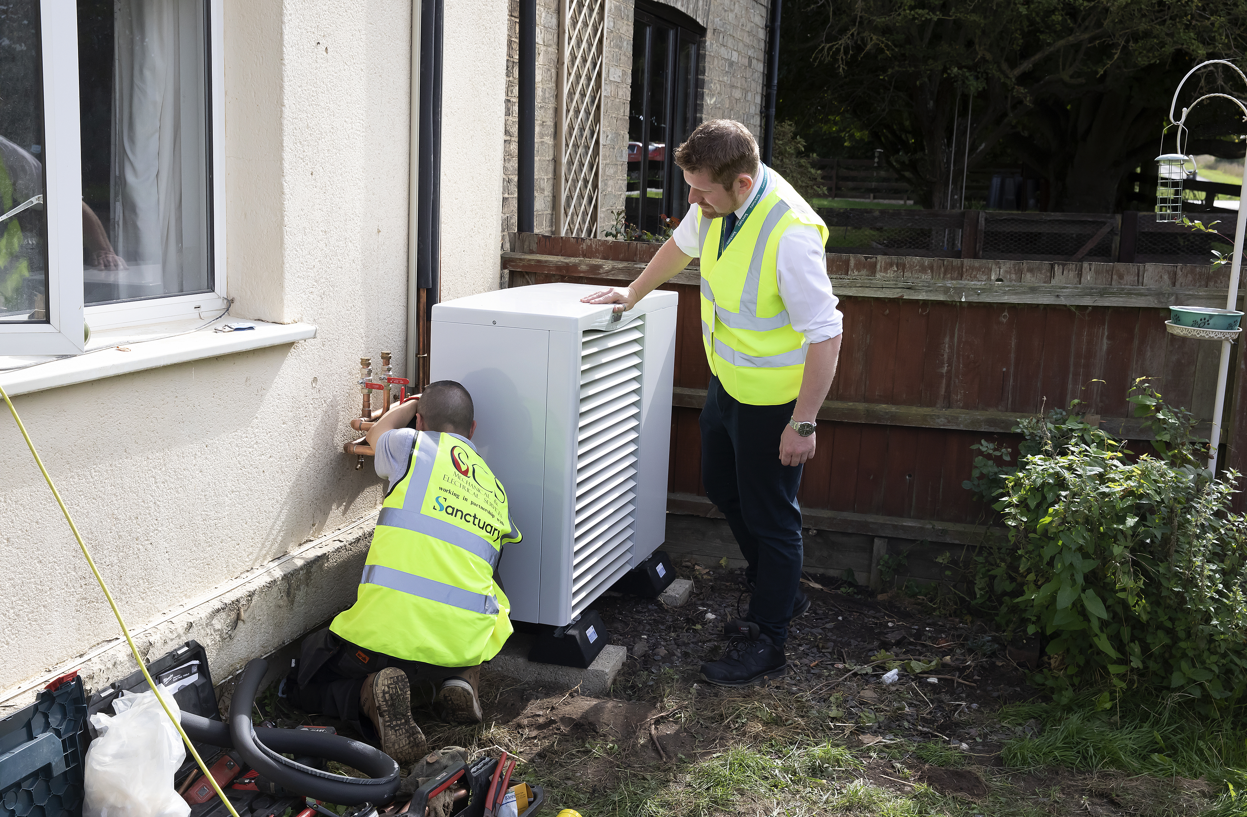 Two white, male Sanctuary staff members wearing high vis jackets are outside a building fitting a Swaffham heat pump
