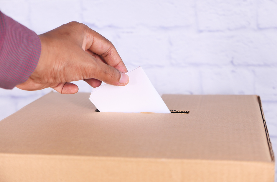 A hand holding a ballot paper posting it into the ballot box below