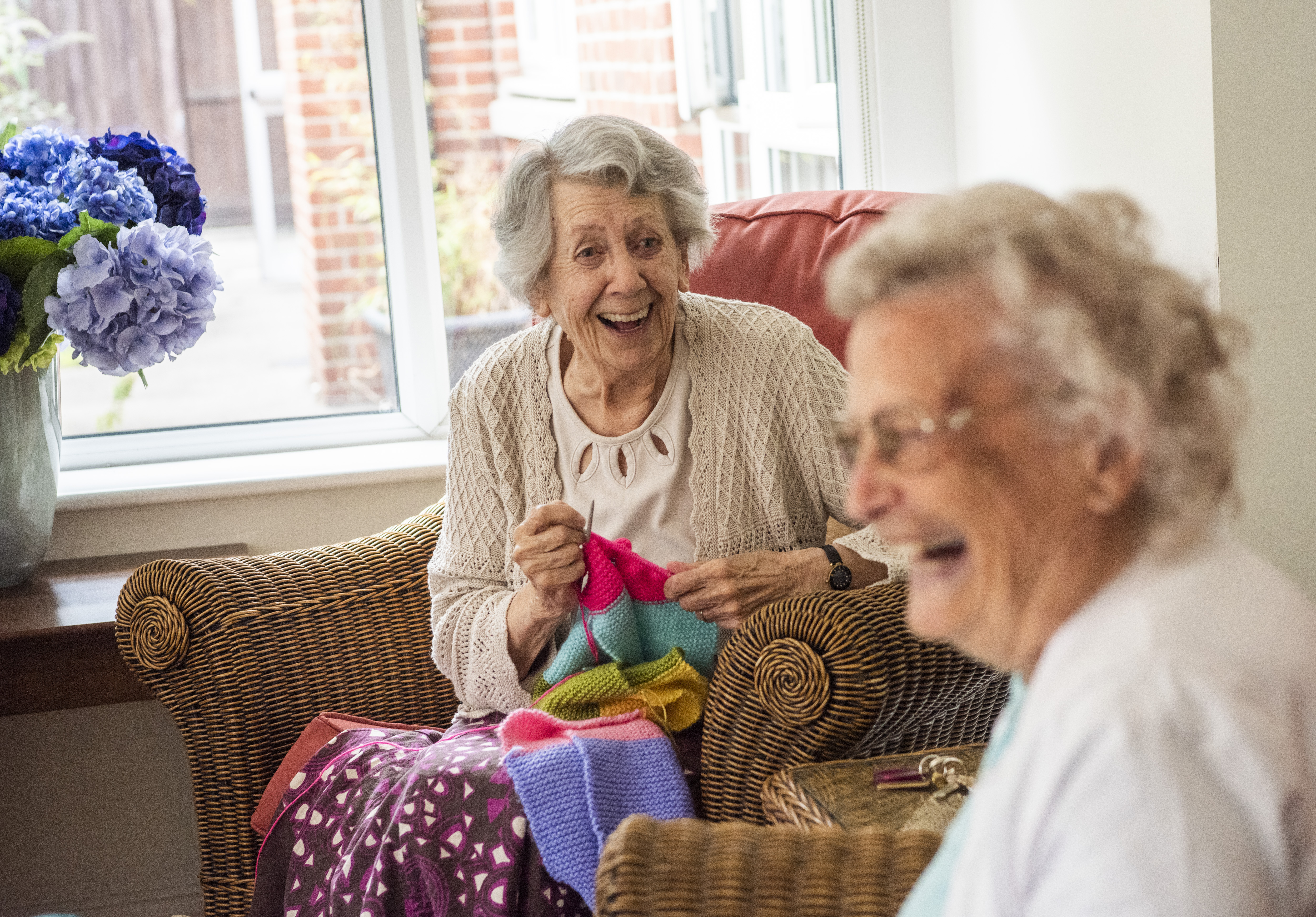 Two elderly women sitting in wicker chairs and laughing whilst knitting with very bright coloured wool