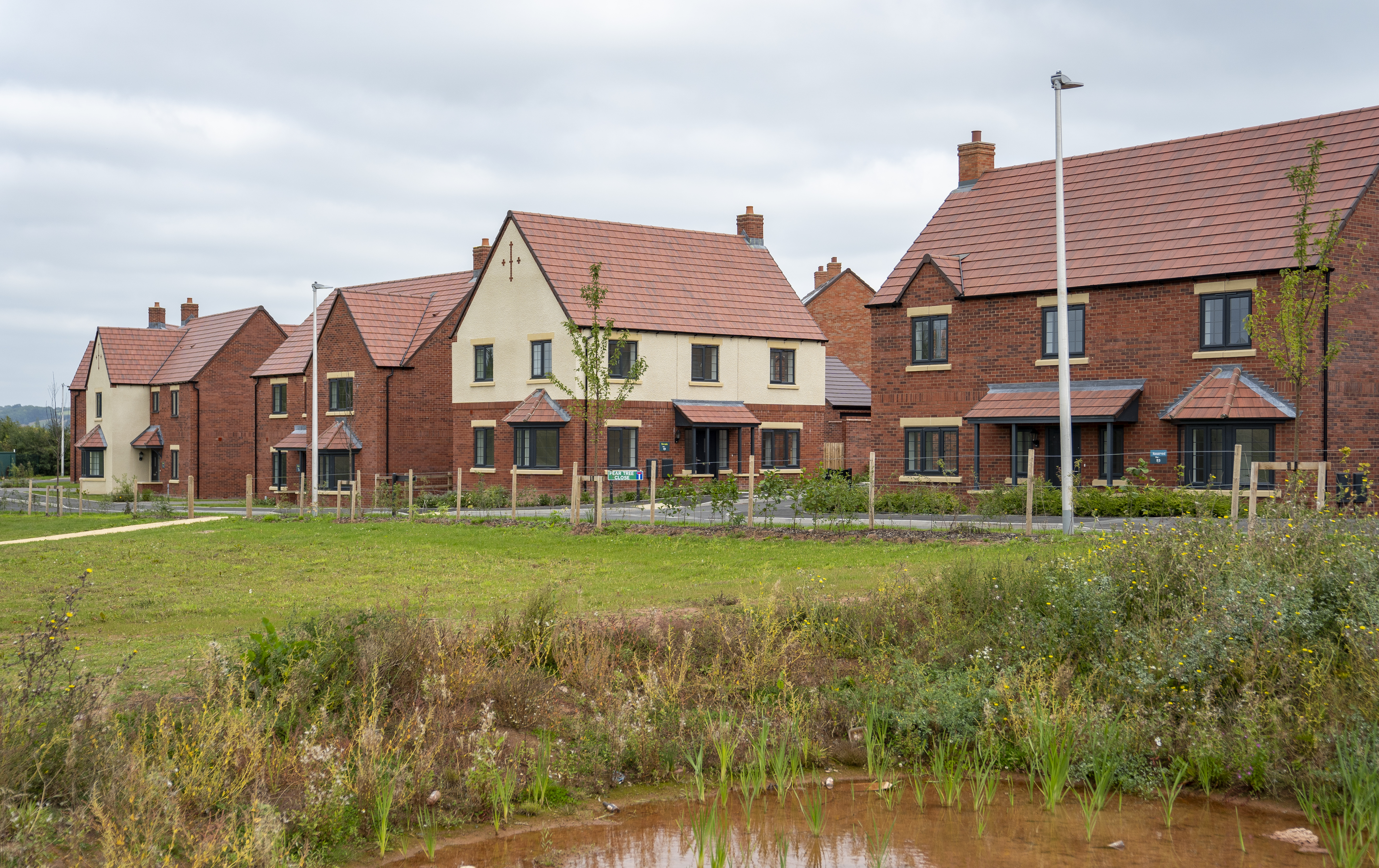 A Sanctuary new-build development estate, build with red brick and cream rendering. There is a large grassy area in the forefront of the image