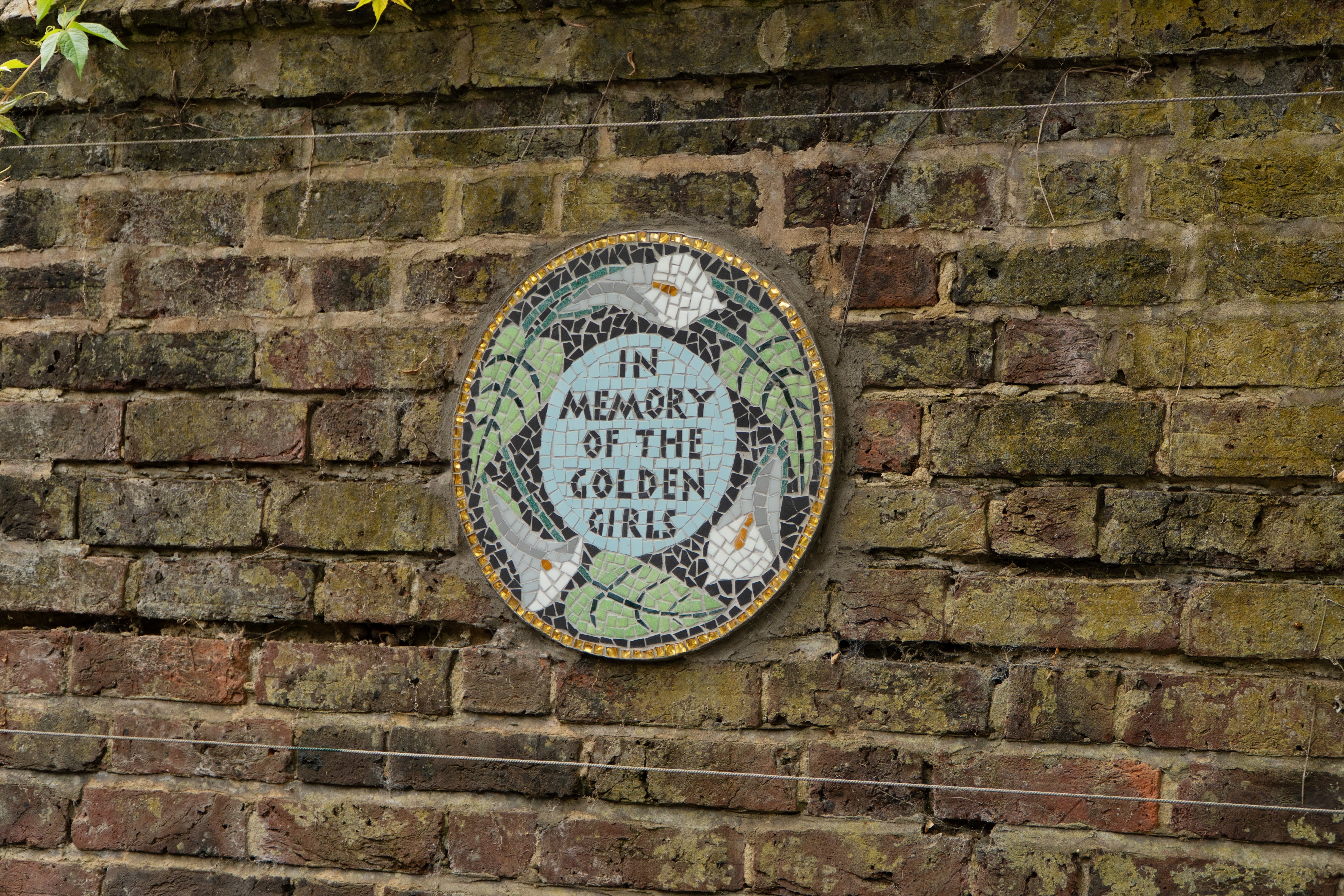 Circular mosaic plaque on a brick wall with the text 'IN MEMORY OF THE GOLDEN GIRLS', surrounded by white birds and green leaves