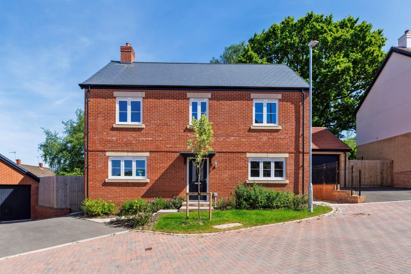Charming two-story red-brick detached home with slate roof, white-framed windows, black front door, front garden with young tree, and driveway leading to garage in a landscaped residential area.