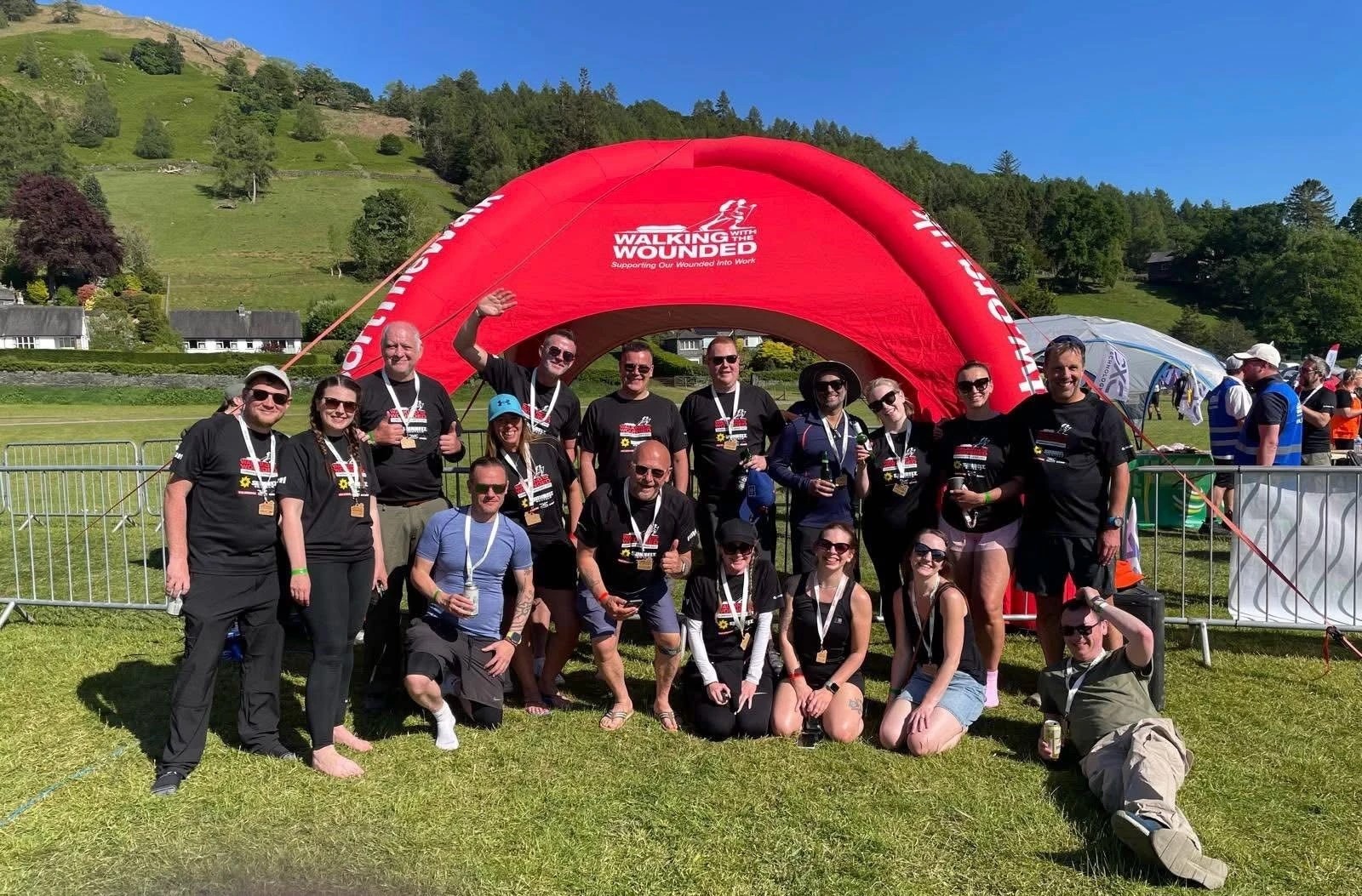 A group of people wearing black 'Walking With The Wounded' t-shirts pose in front of a red inflatable arch with the same logo. They are outdoors on grass with hills and trees in the background. Some are standing, kneeling, or sitting, and many are holding medals.