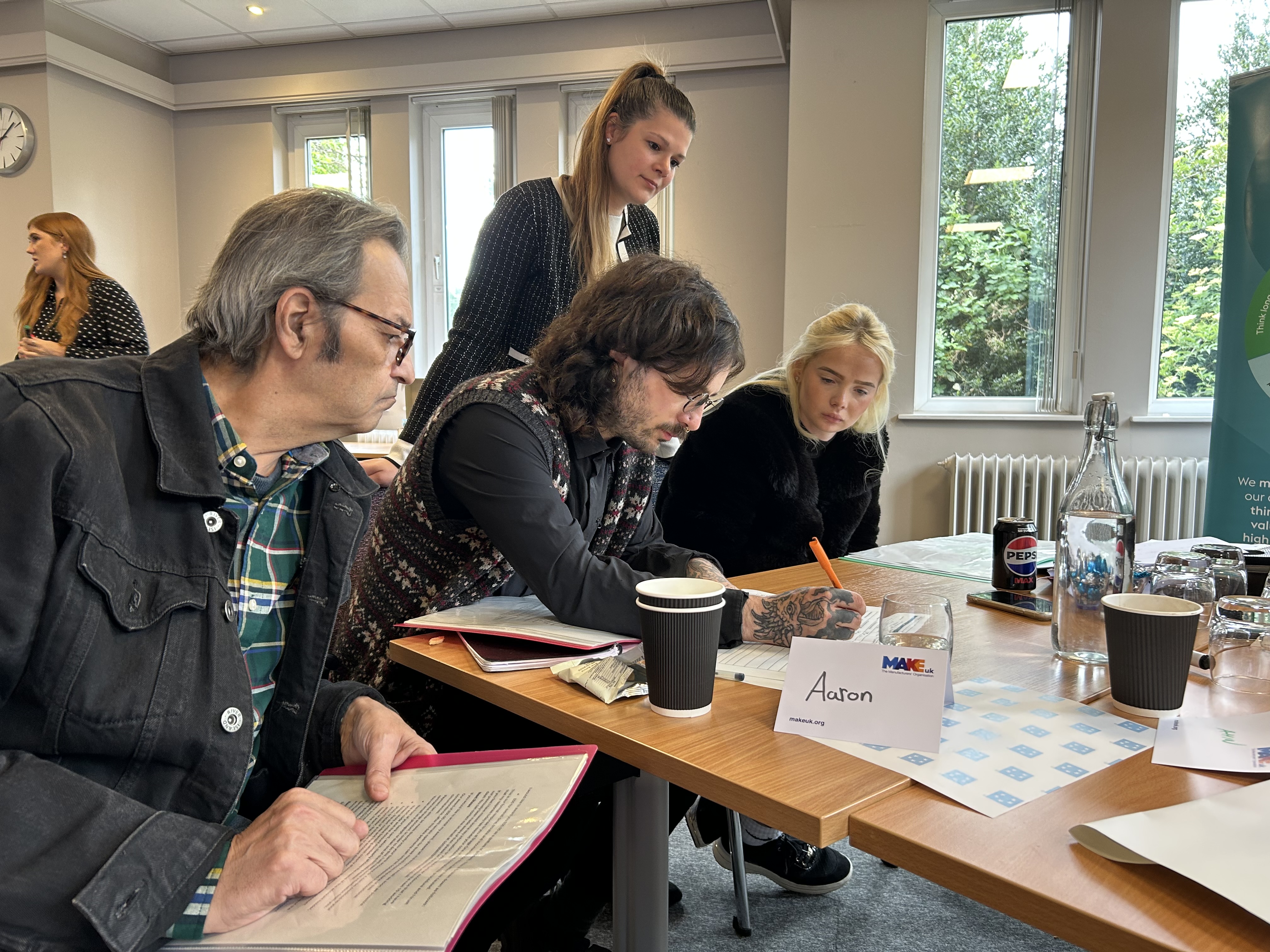 Sanctuary customers seated around a table during a training session on supporting the organisation with complaints. One person stands assisting others. The table has papers, drinks, and a name card reading 'Aaron'. The room is bright with large windows