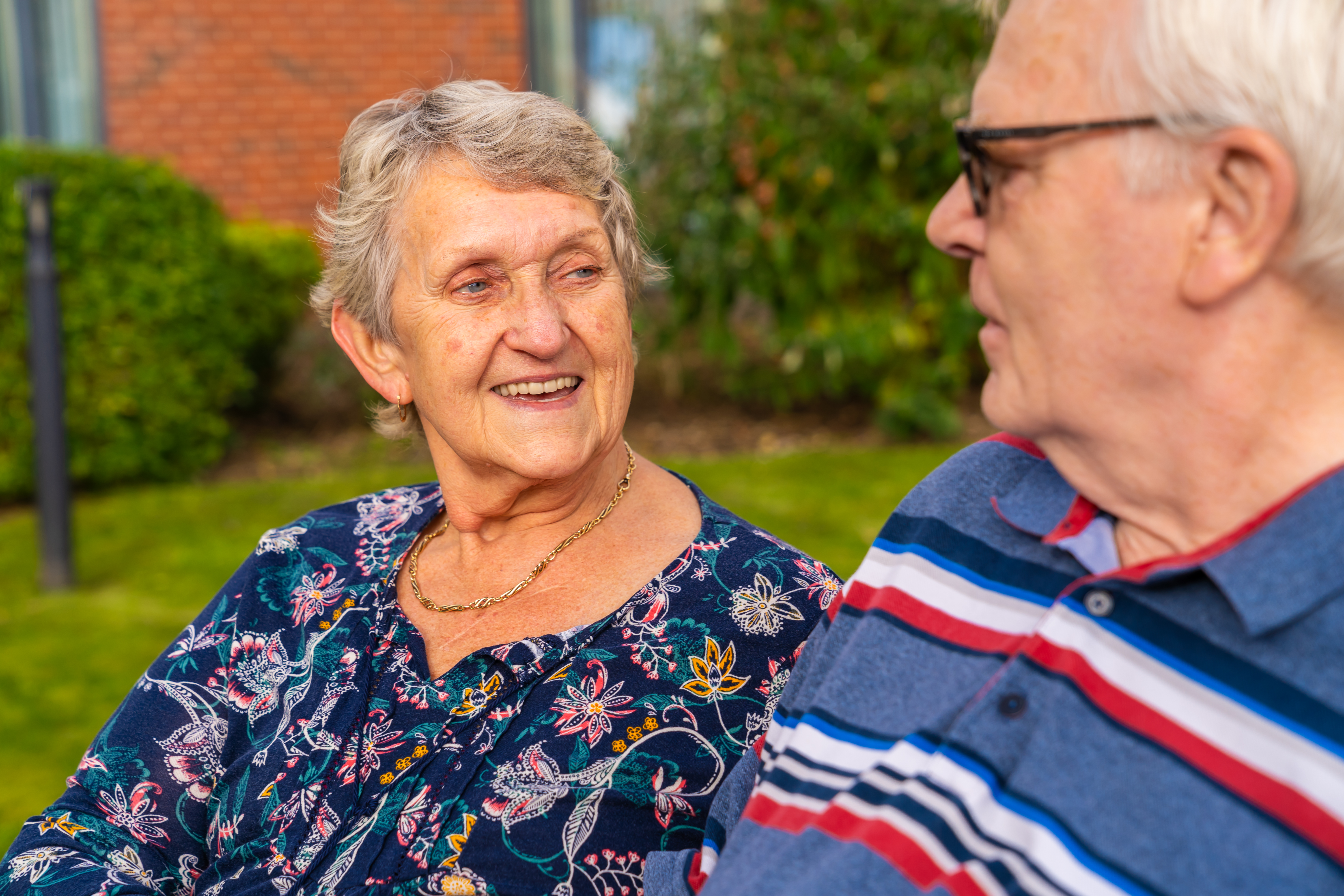 Two elderly individuals are sitting outdoors on a bench, facing each other. One wears a dark floral blouse with a gold necklace, and the other wears a blue polo shirt with red, white, and blue stripes. They appear to be engaged in conversation. The background includes greenery and part of a brick building.