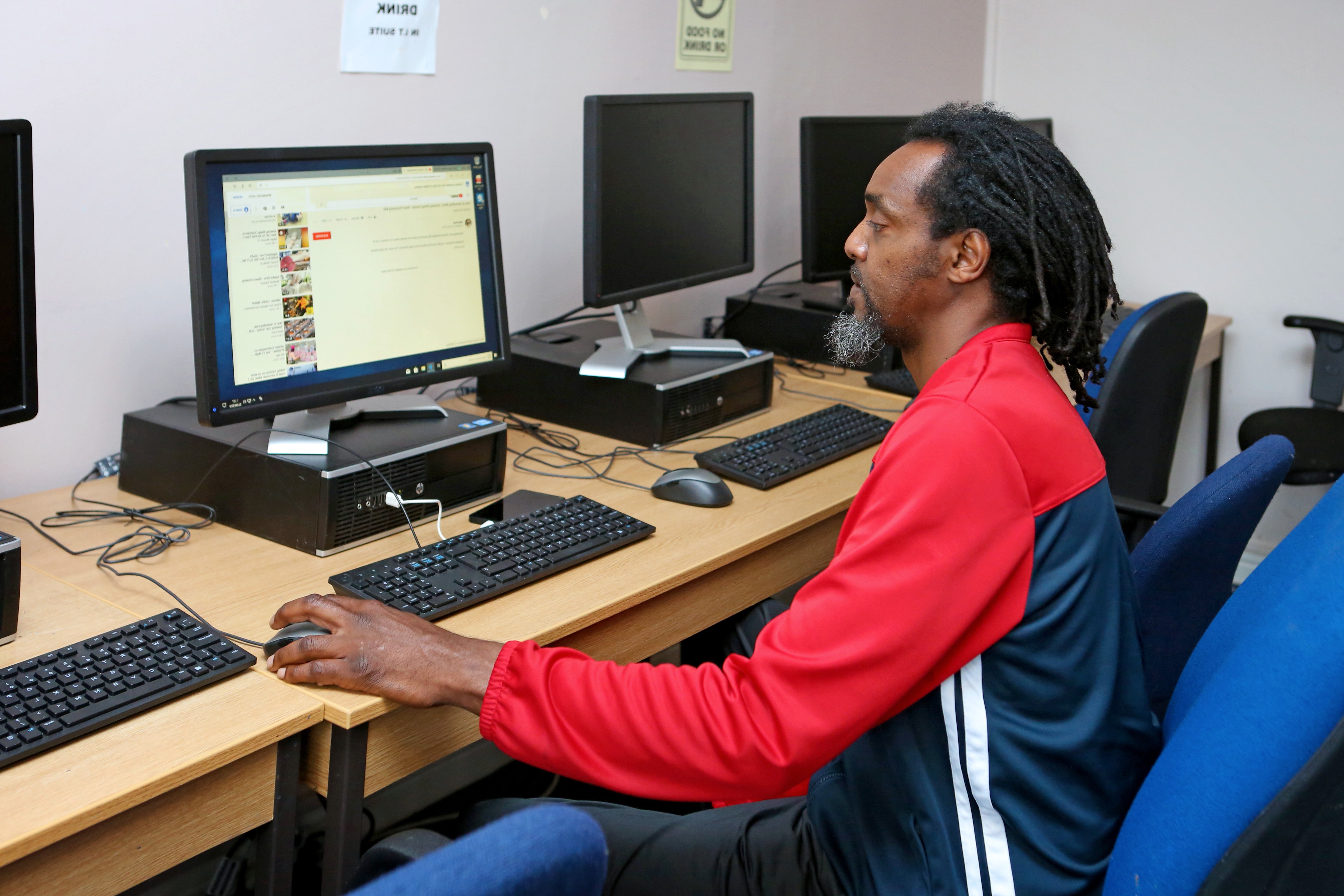 A man with long, curly hair and a beard is sitting at a computer desk in a room with multiple computers. He is wearing a red and black athletic jacket and is focused on the screen in front of him, which displays a web browser. The workspace is simple, with wooden desks and blue chairs.