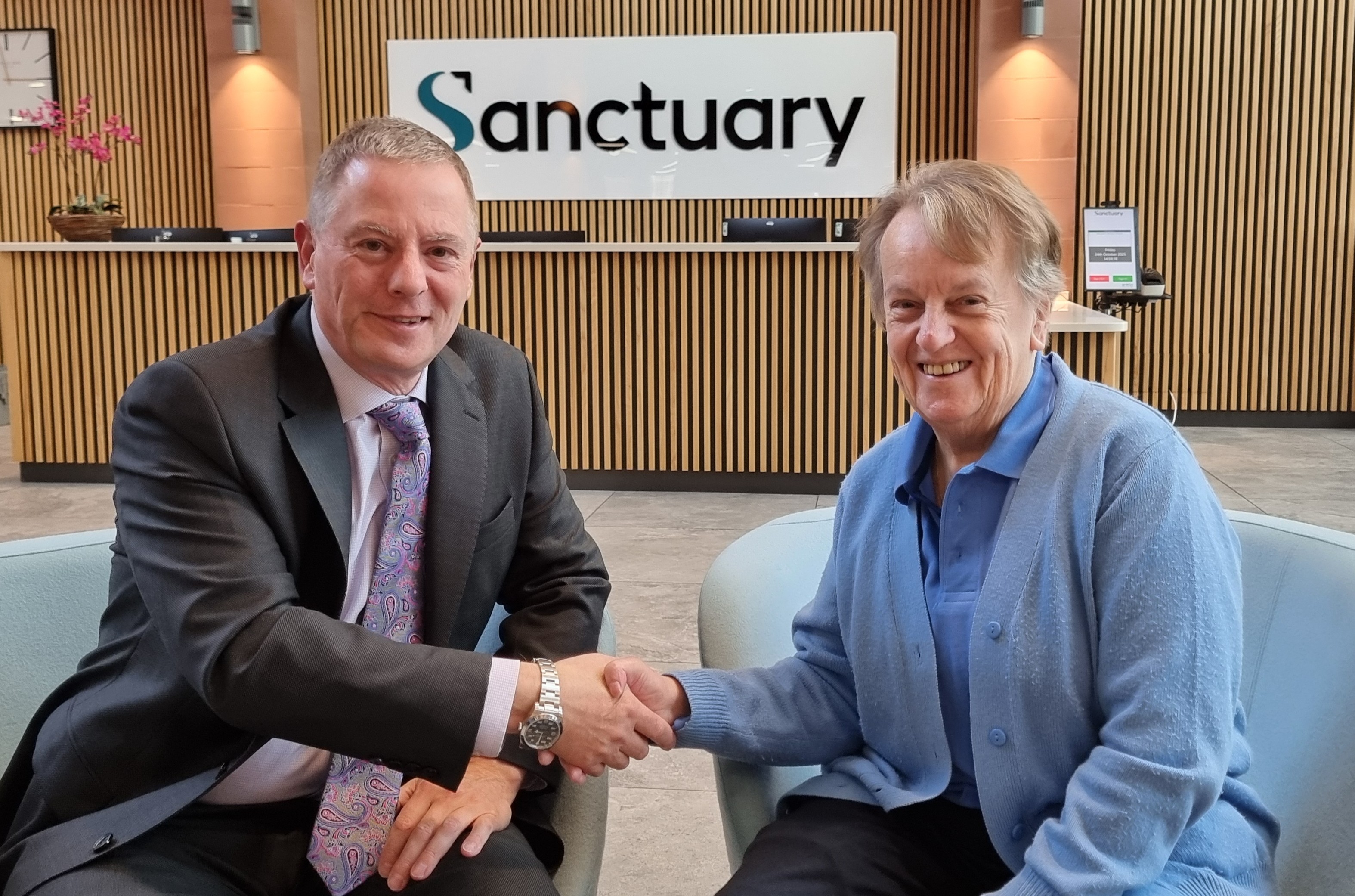 Two people are seated in modern chairs in a reception area, shaking hands. Behind them is a wooden-panelled wall with a large sign that reads “Sanctuary.”