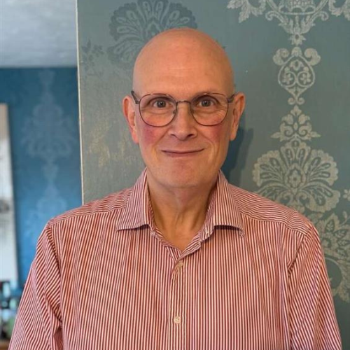 A smiling, bald man wearing round glasses stands against a wall decorated with pale blue wallpaper