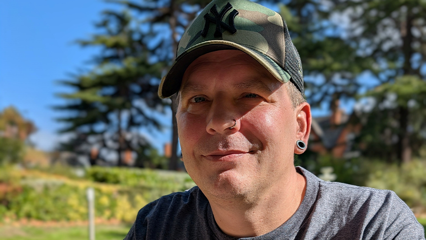 Sanctuary customer Alan Tyla looks towards the camera and smiles, He is sat outdoors in a grassy area and wearing a t-shirt and baseball cap.
