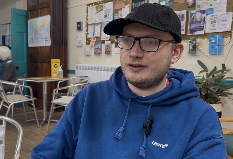Person in blue Levi’s hoodie and black cap sitting in a café with bulletin board and chairs in background.