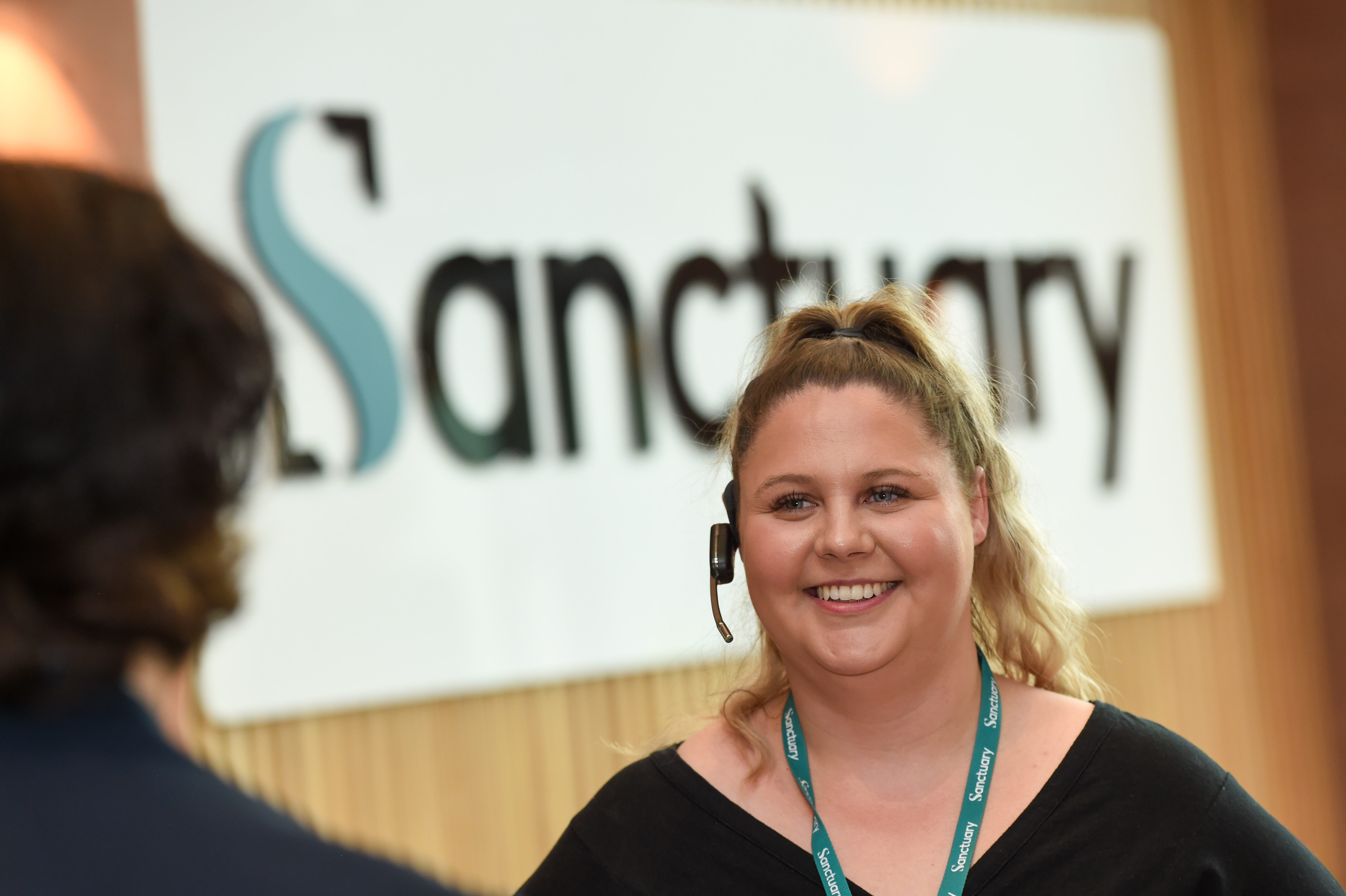 A smiling woman with curly blonde hair is wearing a headset and standing in front of a sign that reads "Sanctuary."