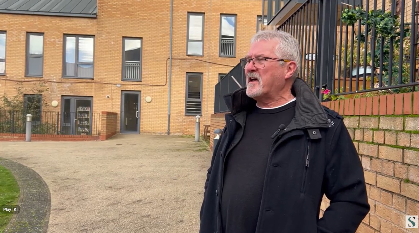 A man with grey hair and beard stands in front of a modern brick block of flats