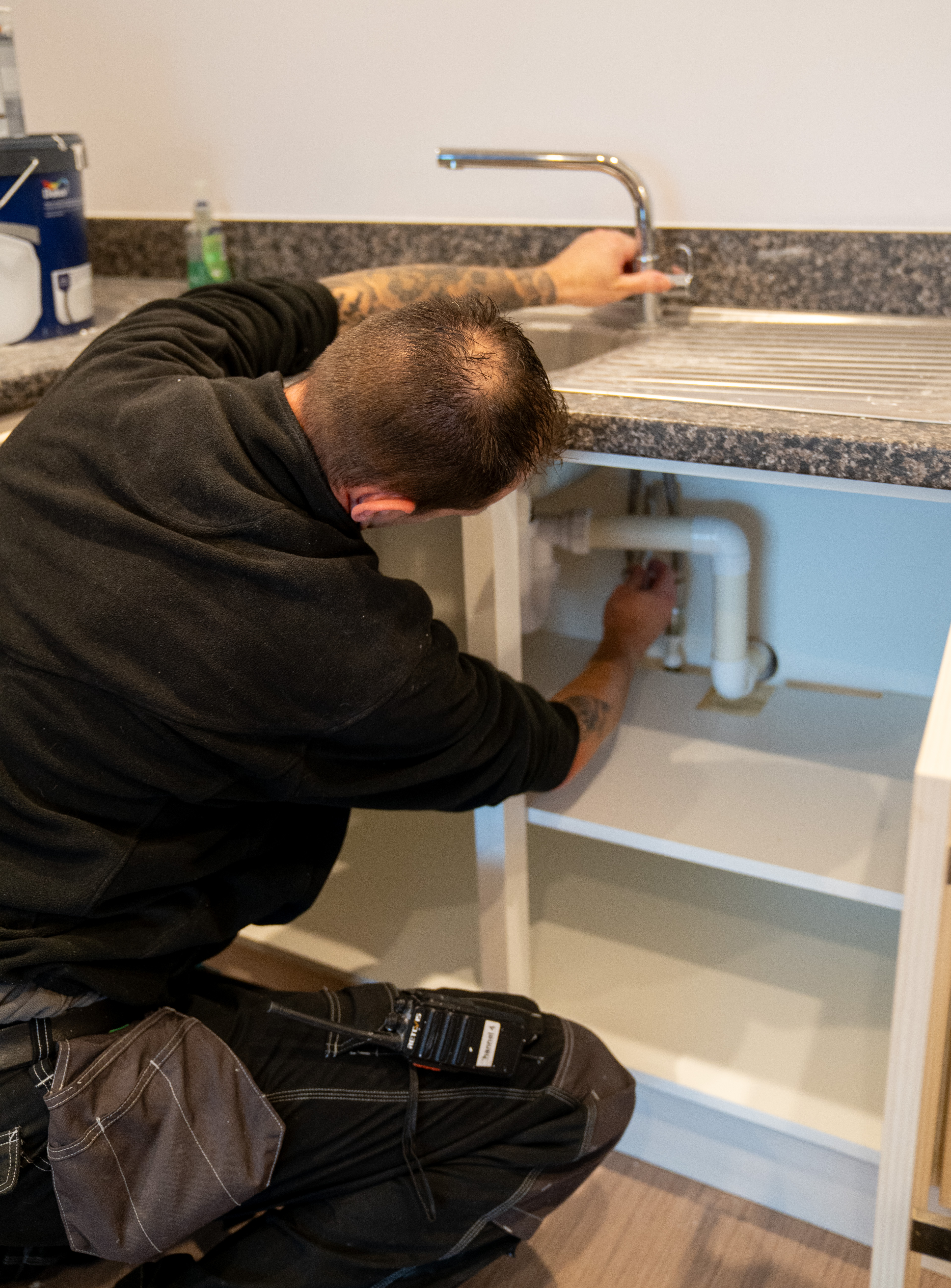 A person wearing a black hoodie and work pants is crouched down under a kitchen sink, working on the plumbing. The sink has a stainless steel surface and a faucet, and there are cleaning supplies and paint cans visible on the countertop.
