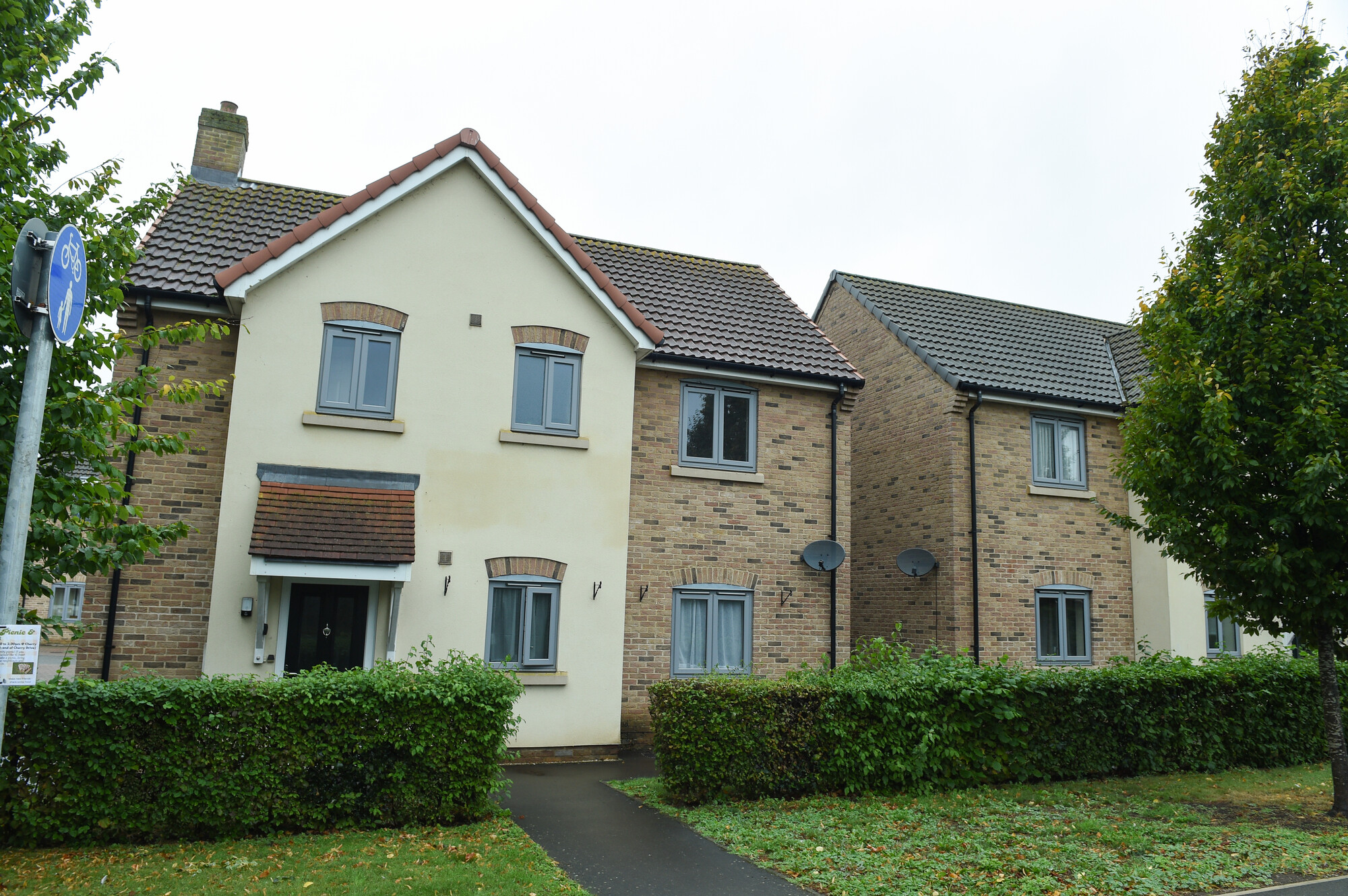 Two modern semi‑detached houses with cream and brick walls, grey windows, hedges and a footpath in front.