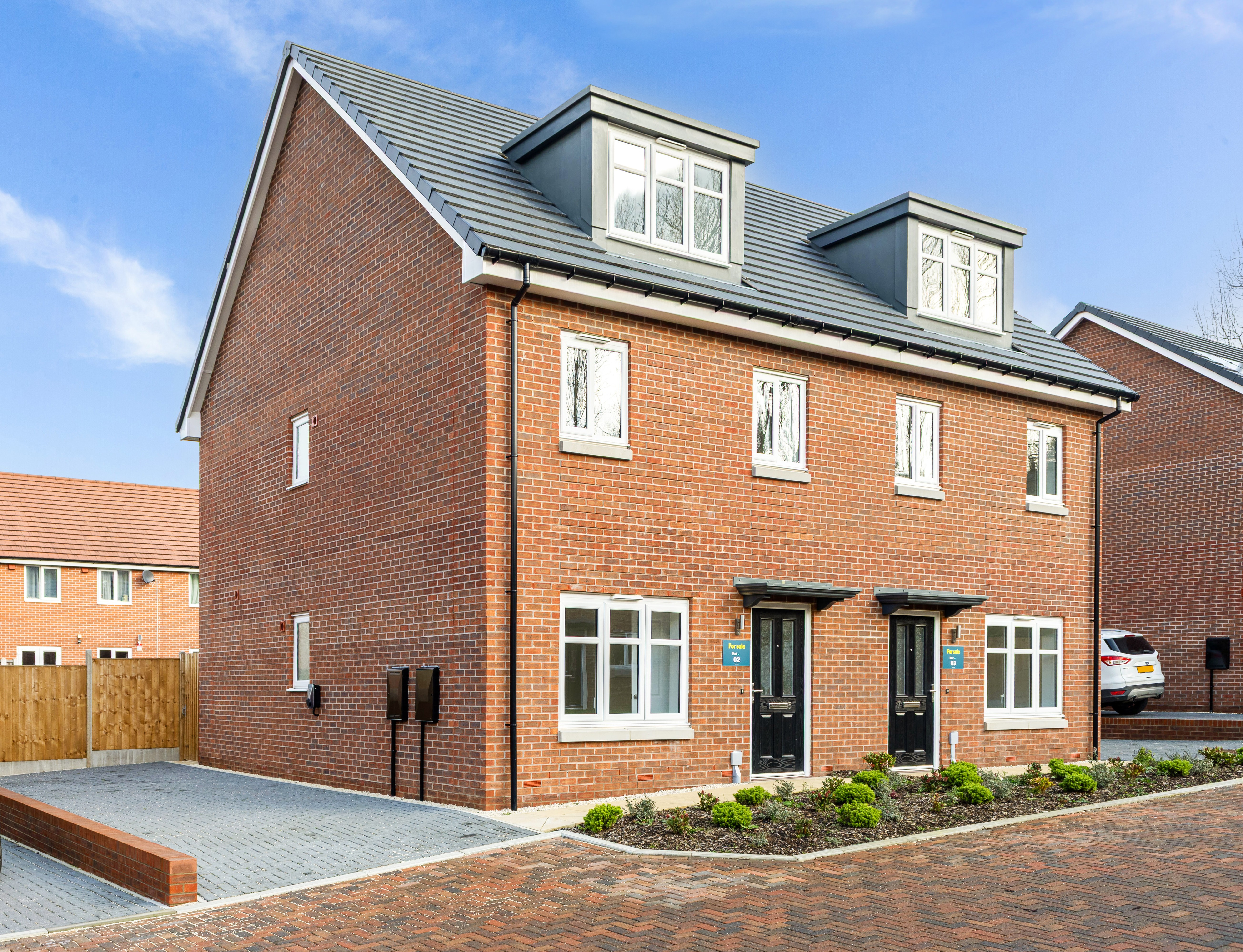 Front view of a red brick semi-detached house with white windows, a black front door and driveway on the left side of the house.