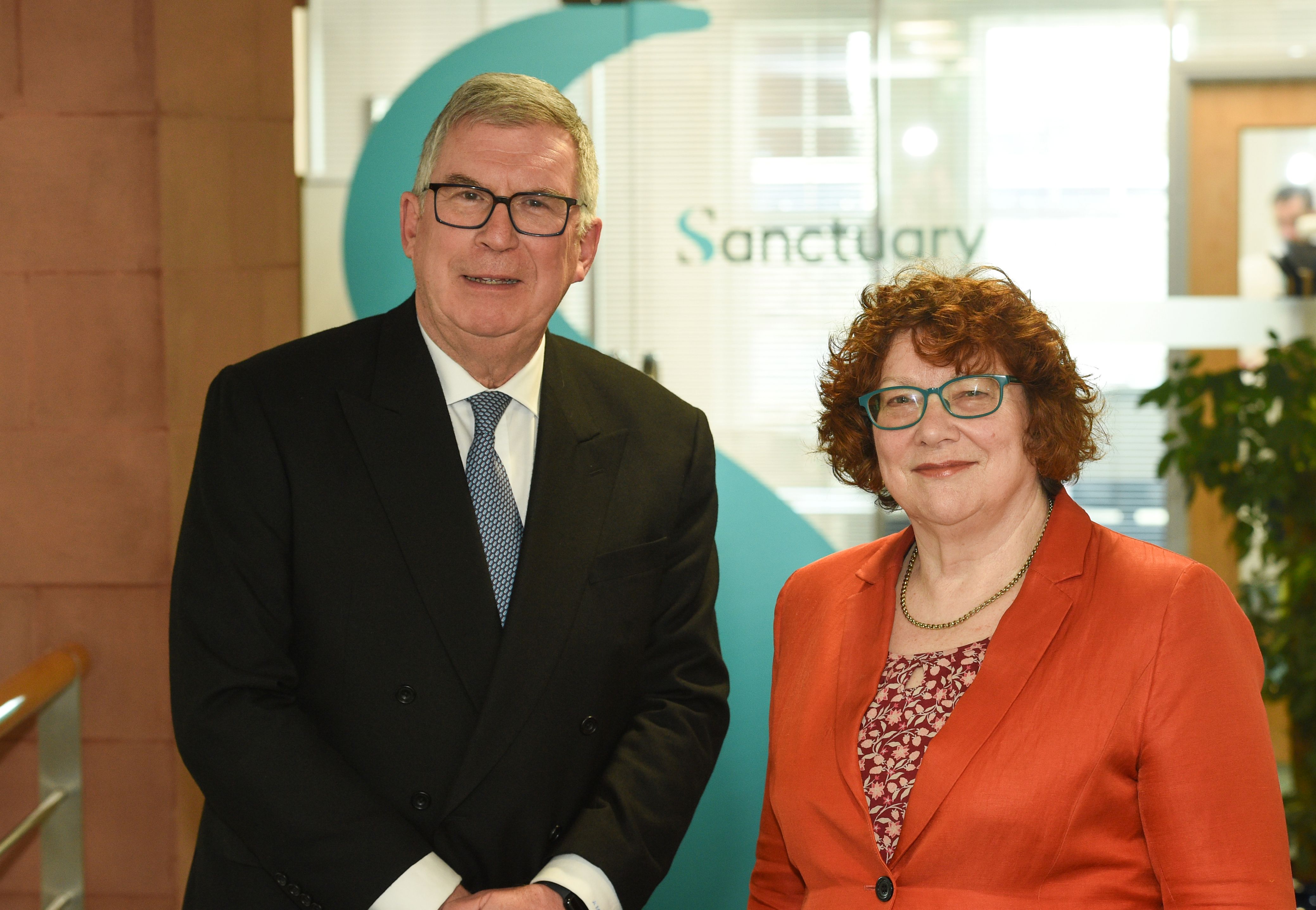Two people standing side by side in an office reception area, with a large Sanctuary logo visible behind them.