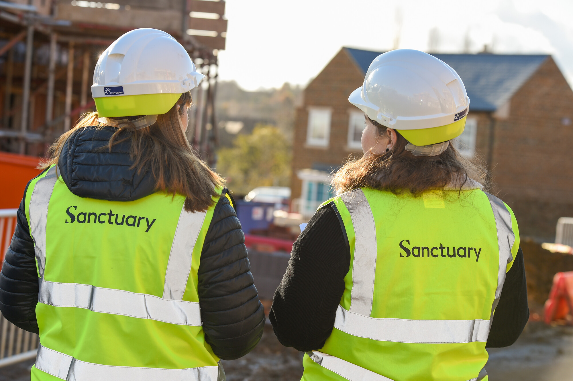 Two women wearing safety helmets and high-visibility vests with the word "Sanctuary" printed on the back are standing with their backs to the camera. They are on a construction site, with scaffolding and partially constructed buildings visible in the background. The scene is illuminated by sunlight, suggesting a clear day.