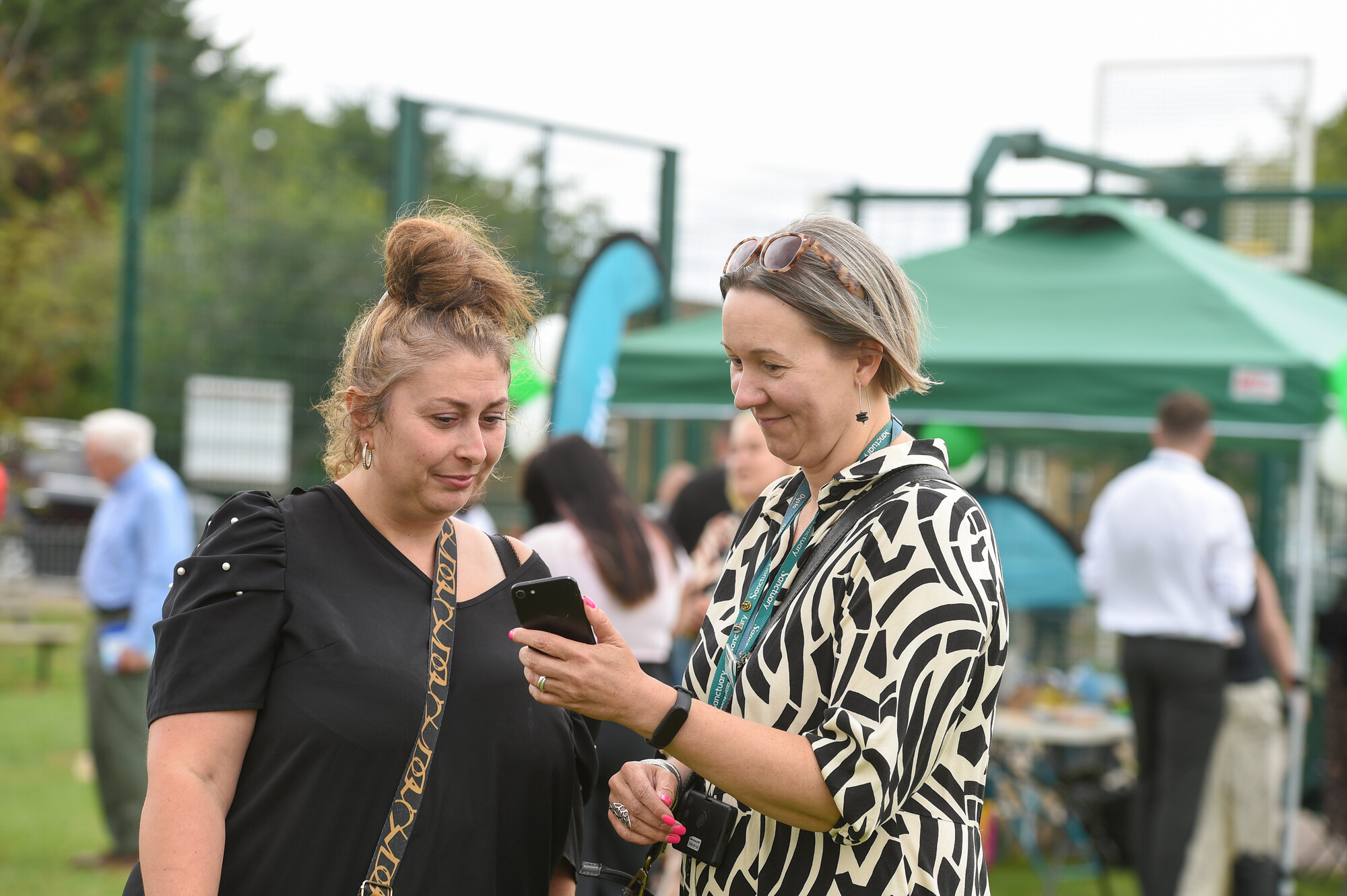 Two women are standing outdoors, engaged in conversation while looking at a smartphone. One woman, with a bun hairstyle and wearing a black top, appears to be listening intently. The other woman, with short grey hair and dressed in a patterned black and white outfit, is showing something on the phone. In the background, there are people mingling and green tents set up, indicating a social event or gathering.