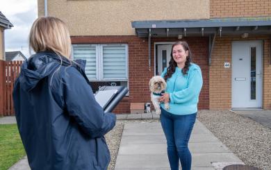 A person outside the front of their home holding a small dog facing another person who looks as though they are working for an organisation with a folder in their hand