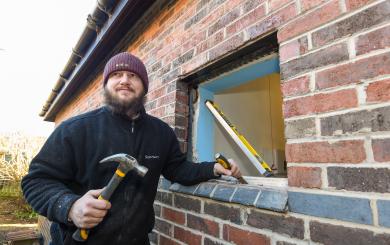 A man in a navy Sanctuary-branded fleece holds a chisel and hammer in front of a window where the double-glazing has been removed