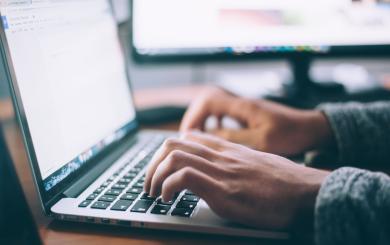 Hands typing on a laptop with a second monitor in the background