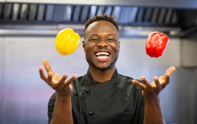 A Sanctuary Care chef wearing chef blacks juggling with a yellow and red pepper whilst laughing