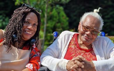 A mother and daughter sat next to each other smiling at the camera, the older woman is sat in a wheelchair