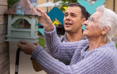 An elderly woman is helped by a younger man to hang a green bird house on a wooden wall