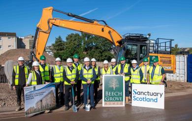 A group of 14 people wearing high vis and hard hats stood in front of a housing development site on a sunny day