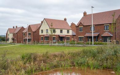 A Sanctuary new-build development estate, build with red brick and cream rendering. There is a large grassy area in the forefront of the image