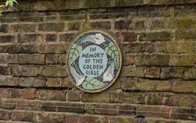 Circular mosaic plaque on a brick wall with the text 'IN MEMORY OF THE GOLDEN GIRLS', surrounded by white birds and green leaves