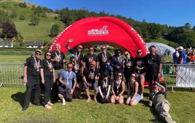 A group of people wearing black 'Walking With The Wounded' t-shirts pose in front of a red inflatable arch with the same logo. They are outdoors on grass with hills and trees in the background. Some are standing, kneeling, or sitting, and many are holding medals.