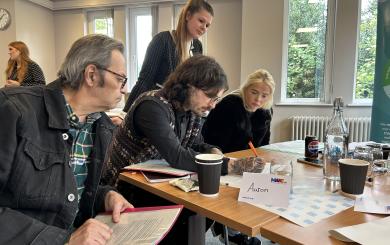 Sanctuary customers seated around a table during a training session on supporting the organisation with complaints. One person stands assisting others. The table has papers, drinks, and a name card reading 'Aaron'. The room is bright with large windows