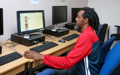 A man with long, curly hair and a beard is sitting at a computer desk in a room with multiple computers. He is wearing a red and black athletic jacket and is focused on the screen in front of him, which displays a web browser. The workspace is simple, with wooden desks and blue chairs.