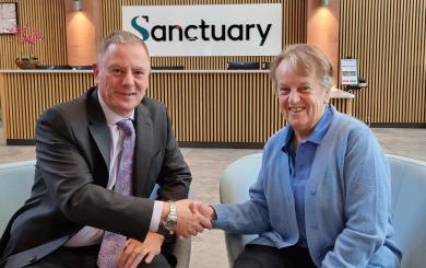 Two people are seated in modern chairs in a reception area, shaking hands. Behind them is a wooden-panelled wall with a large sign that reads “Sanctuary.”