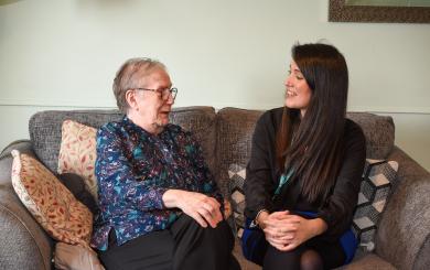 senior woman and a younger woman are sitting together on a cozy gray sofa, smiling and engaging in conversation. The older woman, wearing glasses and a patterned blouse, appears joyful, while the younger woman, dressed in a black top, is laughing. Decorative pillows are arranged on the sofa, and the background features soft lighting and a hint of wall decor, creating a warm and inviting atmosphere.
