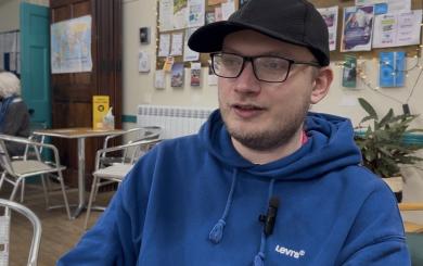 Person in blue Levi’s hoodie and black cap sitting in a café with bulletin board and chairs in background.
