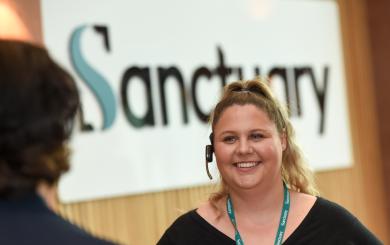 A smiling woman with curly blonde hair is wearing a headset and standing in front of a sign that reads "Sanctuary."