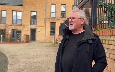 A man with grey hair and beard stands in front of a modern brick block of flats