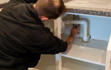 A person wearing a black hoodie and work pants is crouched down under a kitchen sink, working on the plumbing. The sink has a stainless steel surface and a faucet, and there are cleaning supplies and paint cans visible on the countertop.