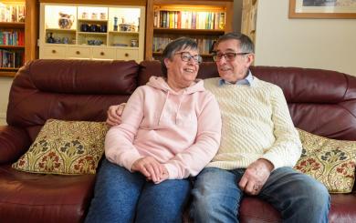 A middle-aged couple sits closely together on a comfortable sofa, smiling at each other. The woman wears a light pink hoodie and glasses, while the man is dressed in a cream-colored sweater and glasses. They are surrounded by a cozy living room with bookshelves and decorative items in the background. The atmosphere is warm and affectionate.