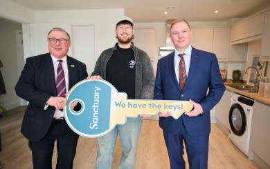 Three men are standing in a modern kitchen. The man in the middle is holding a large, symbolic key with the word "Sanctuary" and the phrase "We have the keys!" printed on it.
