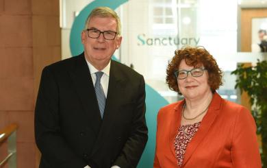 Two people standing side by side in an office reception area, with a large Sanctuary logo visible behind them.