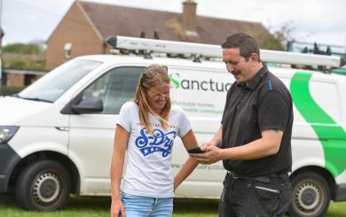 Two people stand outside on a windy day in front of a Sanctuary maintenance van. Sanctuary repairs staff member is showing a resident something on his phone, they are both smiling.