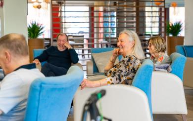 A group of three people is seated in a modern, brightly lit lounge area. In the foreground, a smiling older woman with long gray hair is laughing, wearing a patterned blouse. Beside her, a young girl with short hair is also smiling, while an older man in the background appears to be enjoying a humorous moment. The seating is comfortable, with light blue chairs, and the space is decorated with plants and warm lighting, creating a welcoming atmosphere.