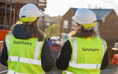 Two women wearing safety helmets and high-visibility vests with the word "Sanctuary" printed on the back are standing with their backs to the camera. They are on a construction site, with scaffolding and partially constructed buildings visible in the background. The scene is illuminated by sunlight, suggesting a clear day.