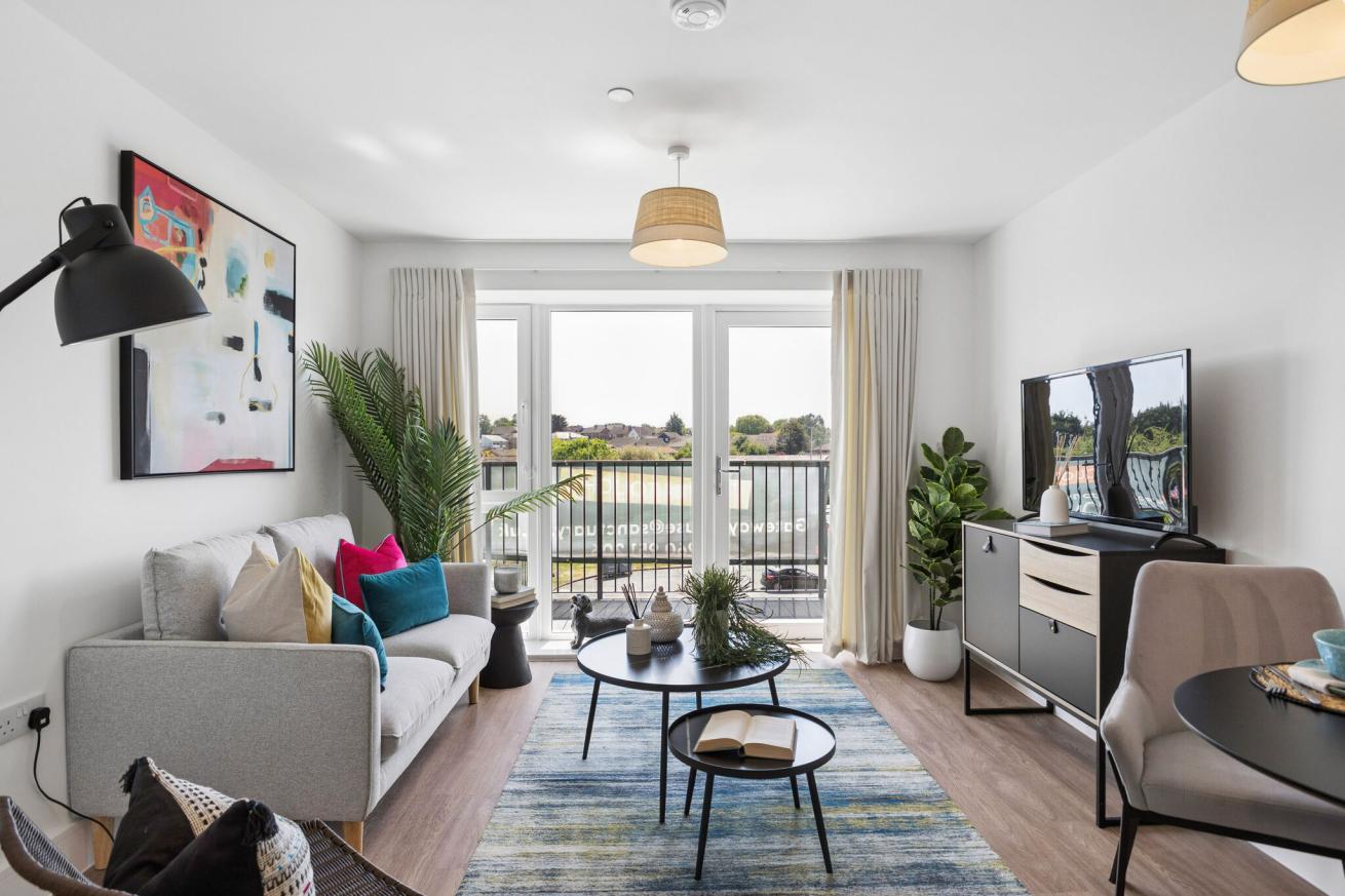 Living room in new build apartment with a light grey sofa with pink, teal and yellow cushions, a tv and stand, 2 small coffee tables and a large abstract piece of artwork above the sofa, and a large decorative plant in the corner