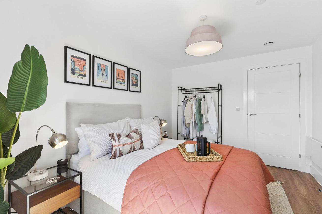 A contemporary bedroom with a double bed with white bedding and a coral-coloured throw, white walls and framed artwork above the bed. There is a clothes rail on one wall, and a bedside table and plant in one corner nearest the bed